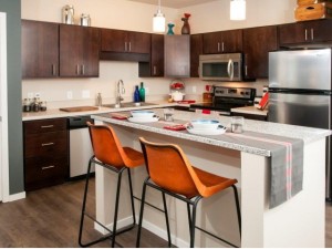 Dark kitchen cabinents along a corner wall with stainless steel appliances. A breakfast bar is set for a meal in the foreground with orange counter stools. Hanging glass lights are over the bar.